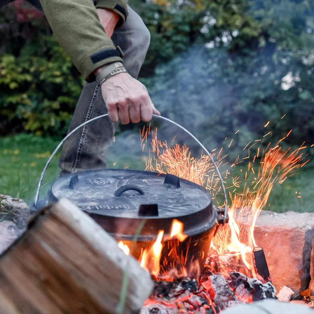 ein schwerer Feuertopf wird am Henkel aus einem heißen Lagerfeuer gehoben